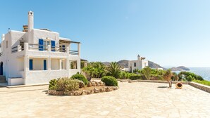 A white building with blue shutters, a stone-paved courtyard, and a view of the sea.