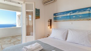 Hotellihuone, jossa on sänky, rantamaiseman kuvaava taulu, avoimesta ovesta avautuva merinäköala ja kivilaattalattia.
