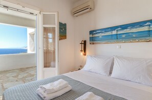 Une chambre d’hôtel avec un lit, un tableau représentant une scène de plage, une vue sur la mer par une porte ouverte et un sol en pierre.
