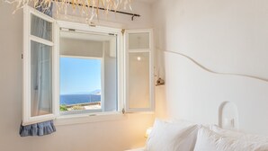 Ein Schlafzimmer mit Bett, einem Fenster mit Vorhängen, einer Nachttischlampe und Blick aufs Meer.
