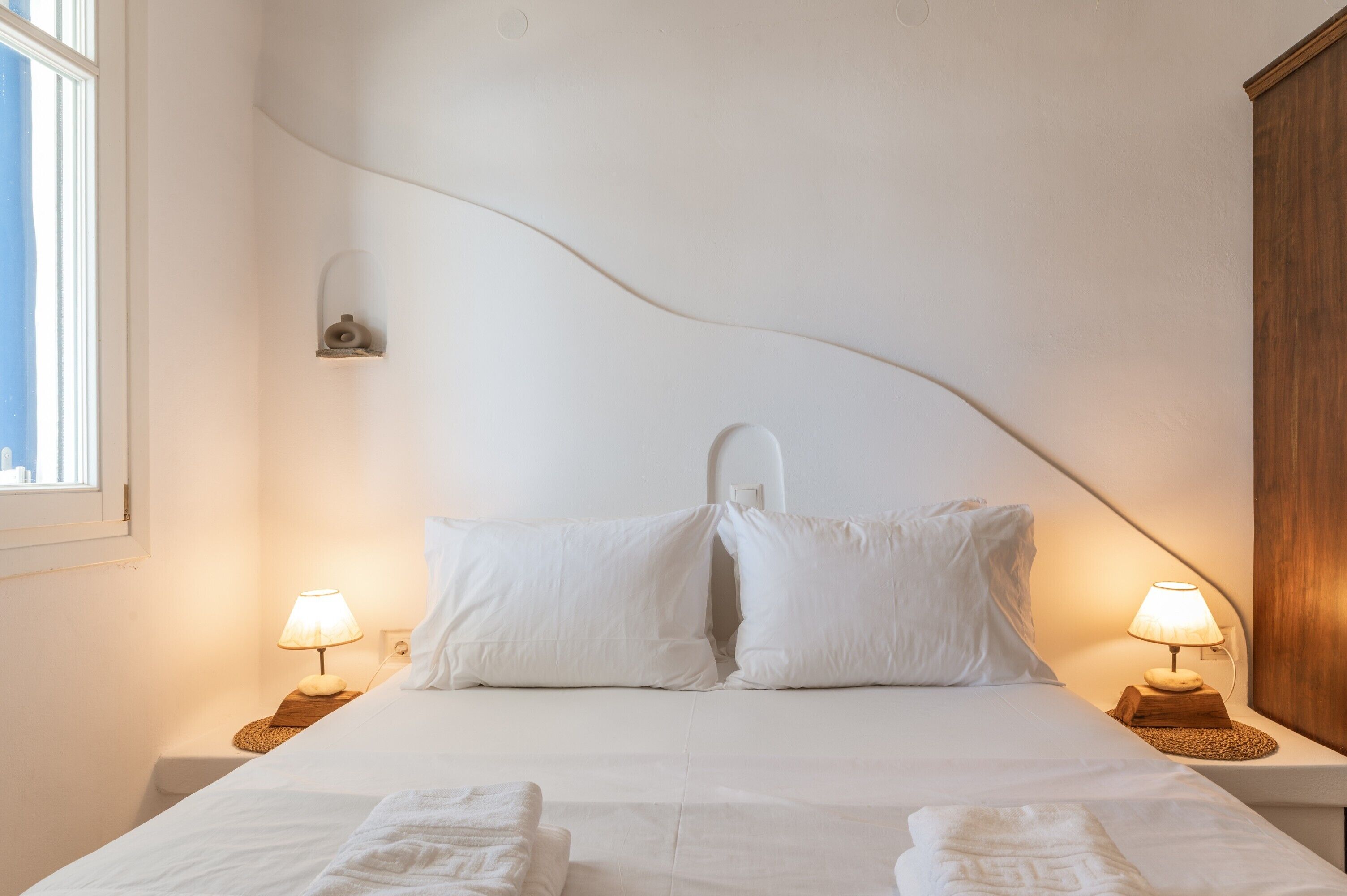 Een net opgemaakt bed met kussens, twee nachtlampjes en een klein plankje met een decoratief object.