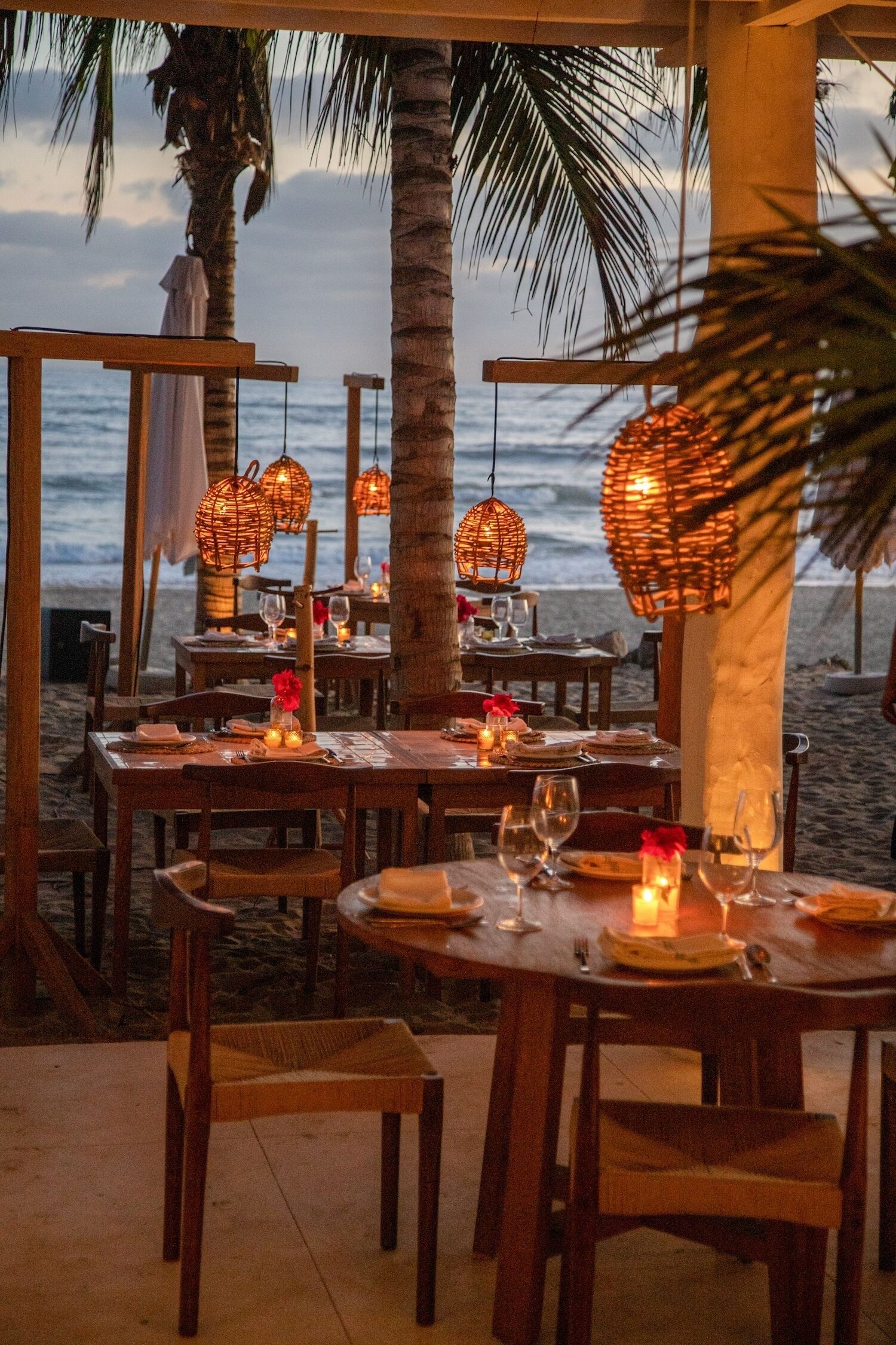 Een restaurant aan het strand met houten tafels en stoelen, tafels met kaarsen en hangende rieten lampen.
