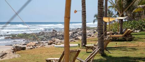 Un área frente a la playa con sillones de madera y una mesita, palmeras y vistas al océano.