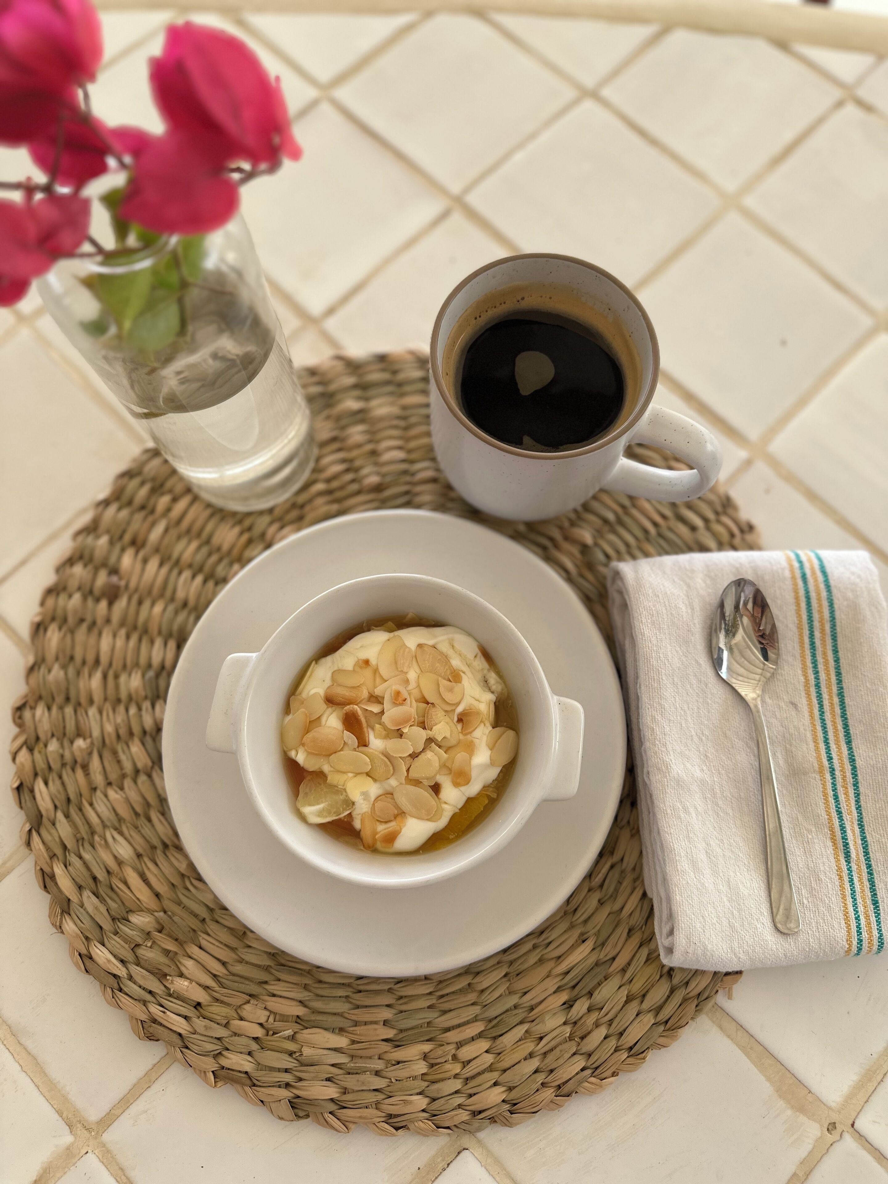 Una mesa dispuesta con una taza de café, un plato de comida cubierto de almendras, una cuchara y una servilleta.