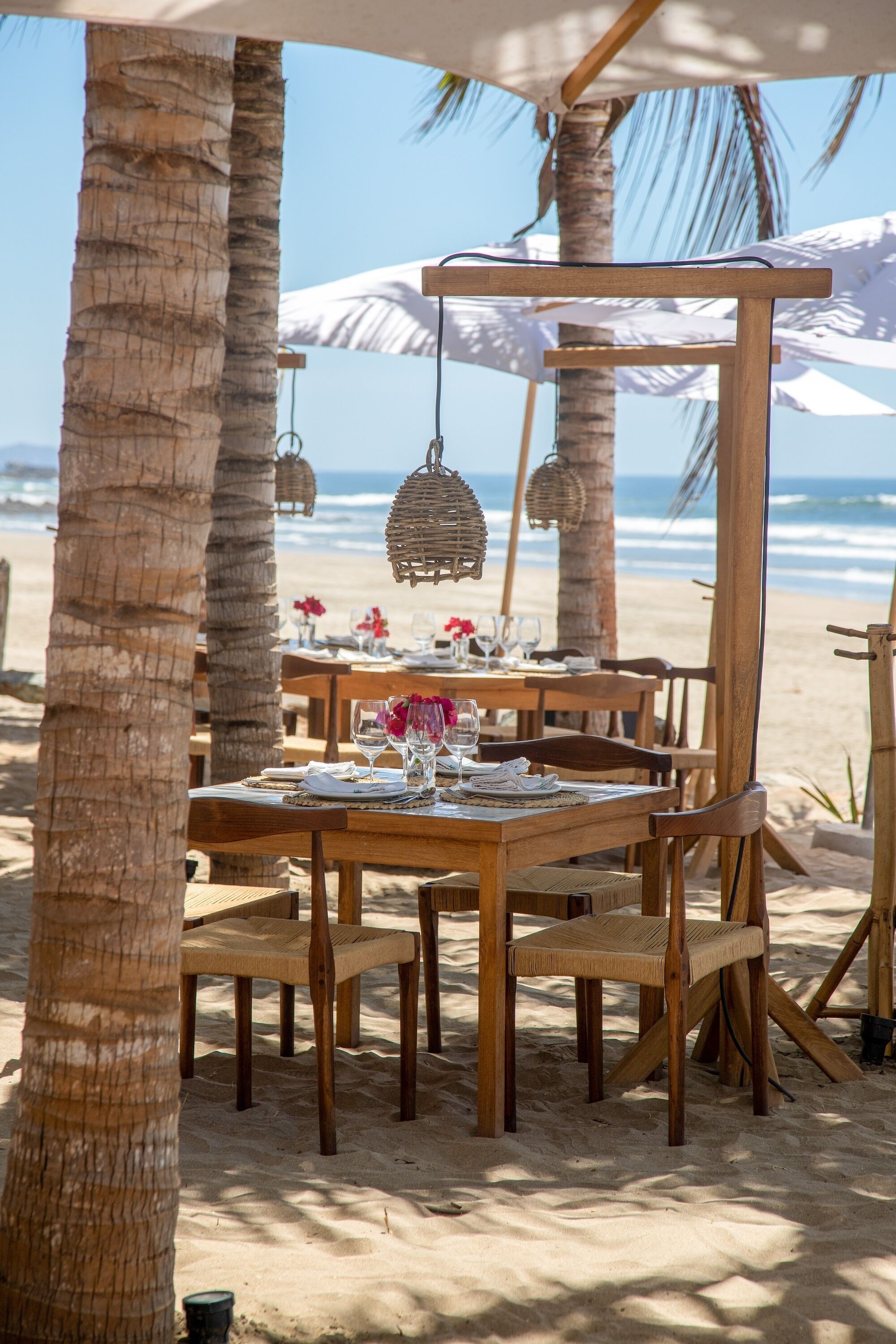 Un conjunto de comedor exterior de madera con mesas y sillas, rodeado de palmeras y vistas a la playa.