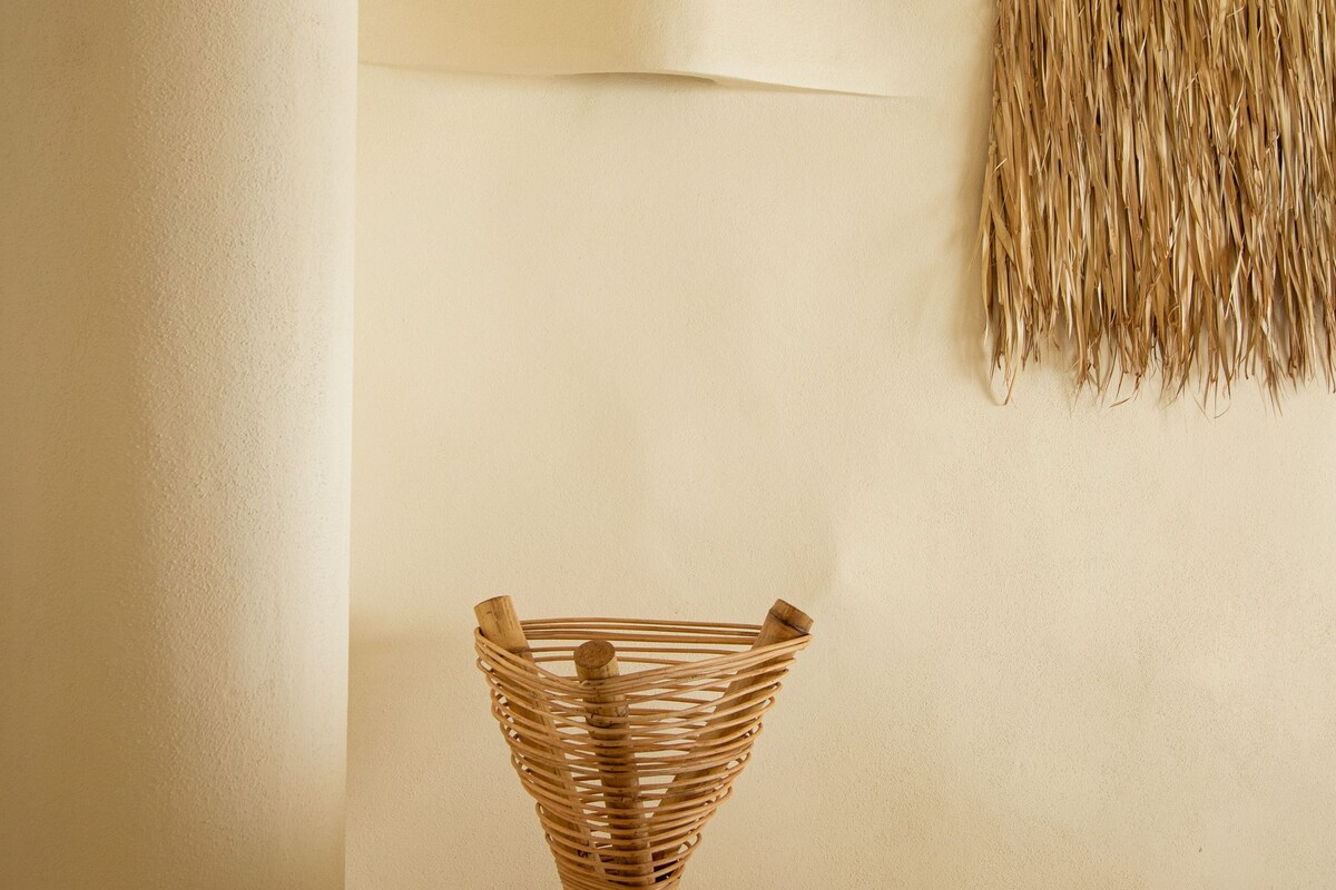 A room with a woven basket on a stand, a wall-mounted hanging, and a plain wall.