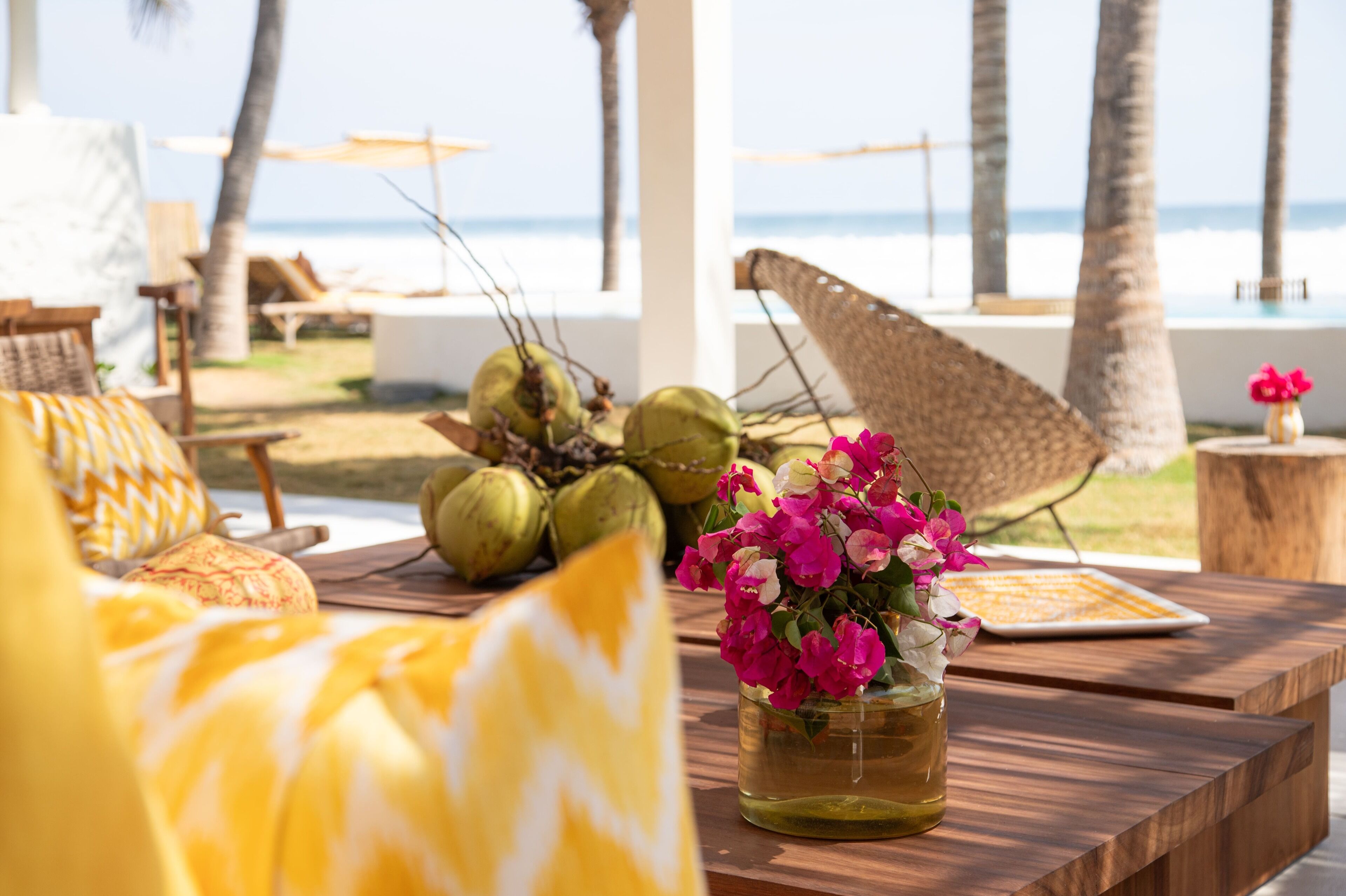 Una mesa de madera con un jarrón de flores y cocos, un paraguas tejido y vistas a la playa y el océano.
