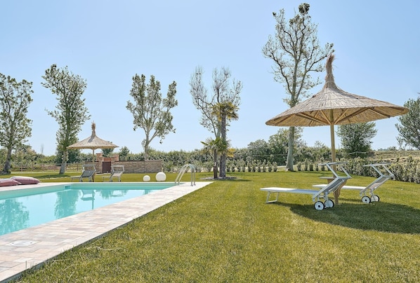Une piscine entourée de chaises longues et de parasols, elle-même bordée d’une pelouse bien entretenue et d’arbres.