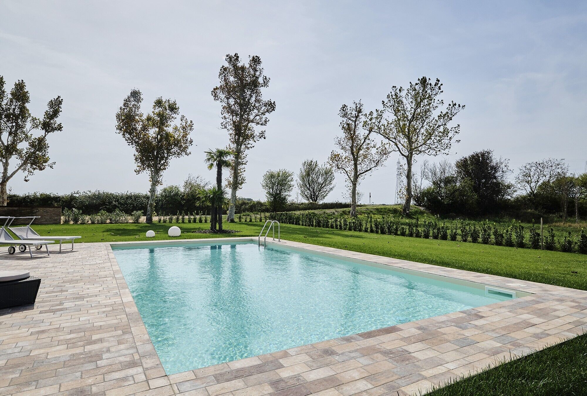 Une piscine rectangulaire bordée d’une terrasse carrelée, entourée d’une pelouse bien entretenue et d’arbres.