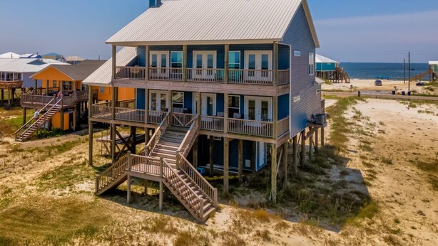 A two-story beachfront house with a wooden deck and stairs leading to the upper level.
