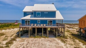 A two-story house with a balcony, wooden deck, and a sandy beach in the background.