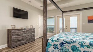 A bedroom with a bed, a wooden dresser, a flat-screen TV, and a balcony with a view of the ocean.