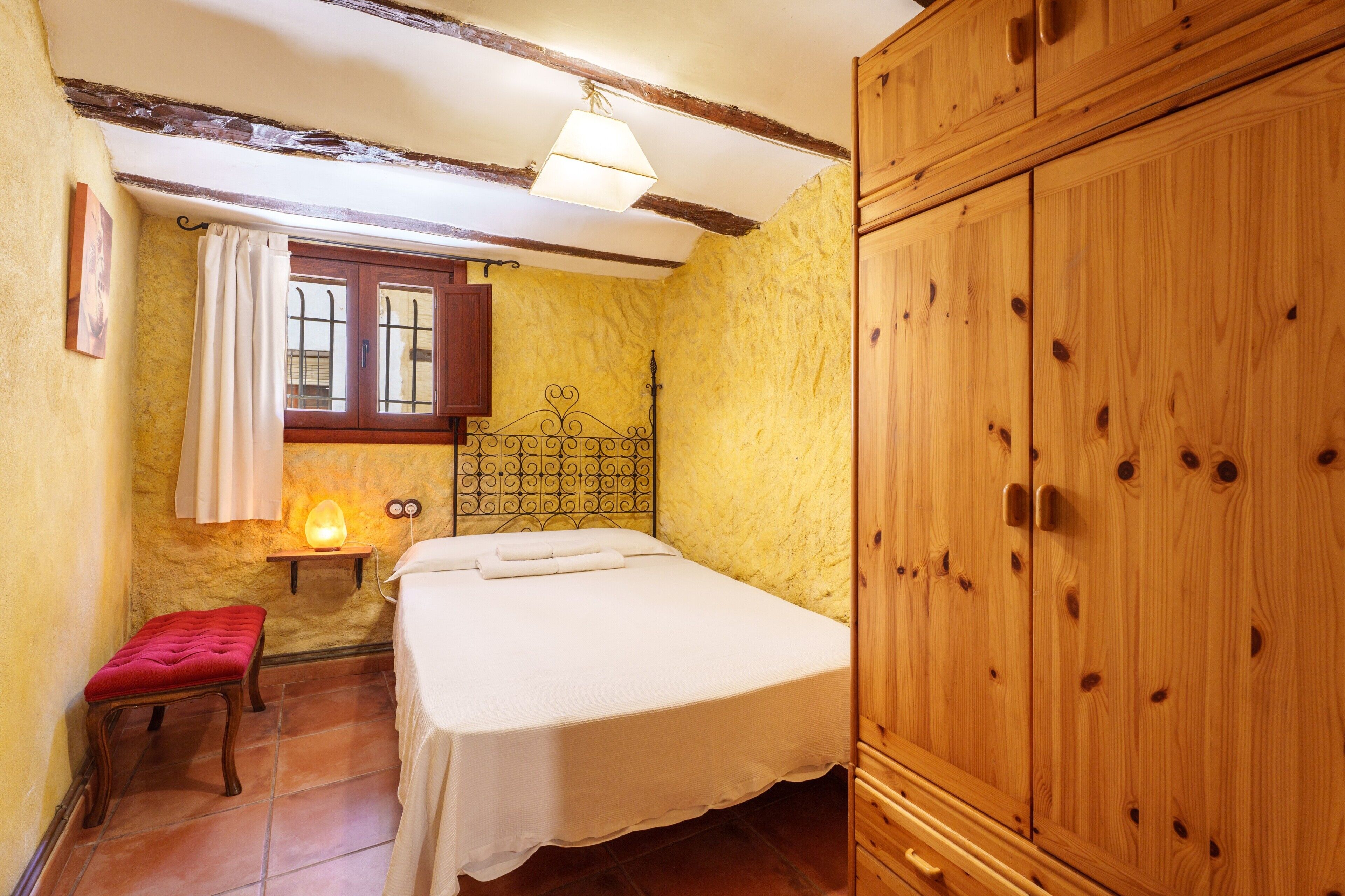 Un dormitorio con cama, un armario de madera, una ventana con cortinas, una mesita de noche con lámpara y un banco rojo.
