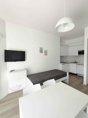 Ein modernes, kompaktes Wohnzimmer mit einer Küchenzeile, einem an der Wand montierten Flachbildfernseher, einem Bett mit grauem Matratze sowie einem Essbereich mit weißem Tisch und Stühlen.