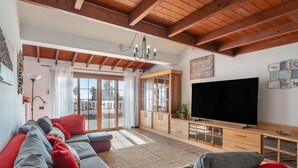Amplio salón con televisor grande, vigas de madera y puerta corredera de cristal que da a un área exterior.