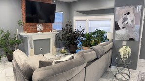 A living room with a grey sofa, a TV mounted on a brick wall, and various potted plants. - Luxury Fully Furnished Spacious 3 BDR 3 Full BTH Columbus Home with Amenities! (Columbus)