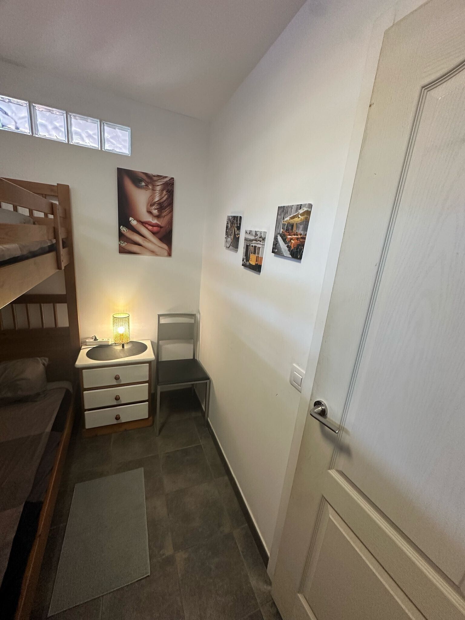 Een kleine, praktische kamer met een stapelbed, een nachtkastje met lamp, een stoel en een deur.