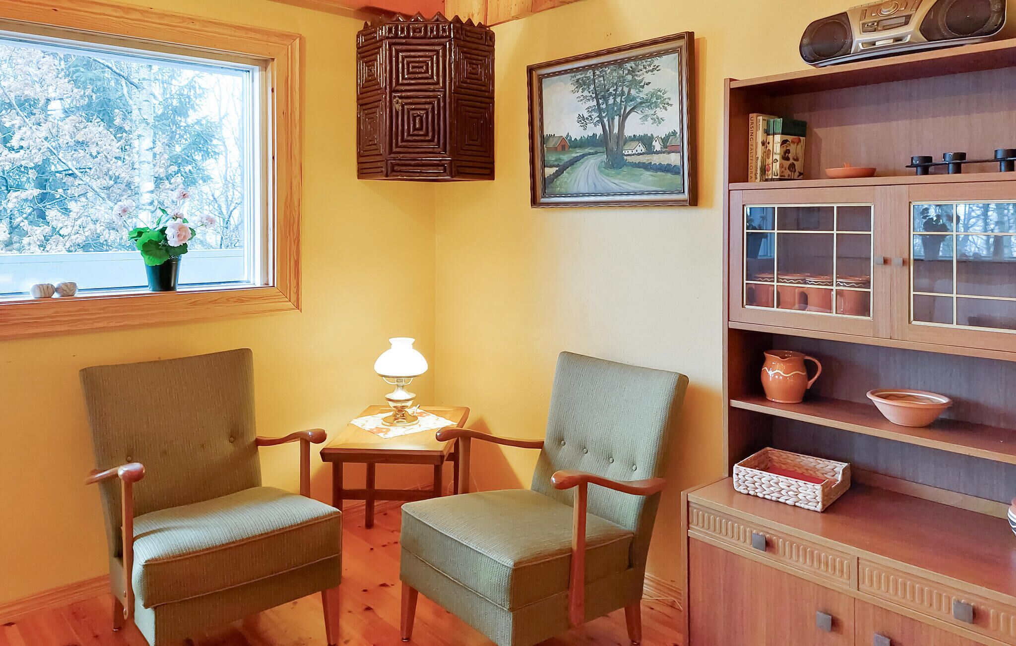 A cozy room with two upholstered chairs, a small table with a lamp, a wooden cabinet with shelves, and a window with a view of trees.