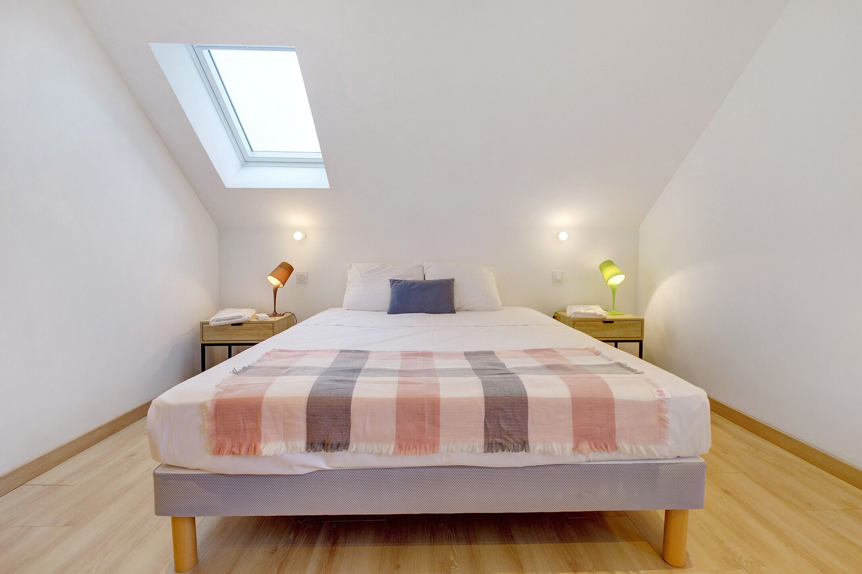 A bedroom with a skylight, a bed with a checkered blanket, bedside lamps, and a nightstand with a lamp.