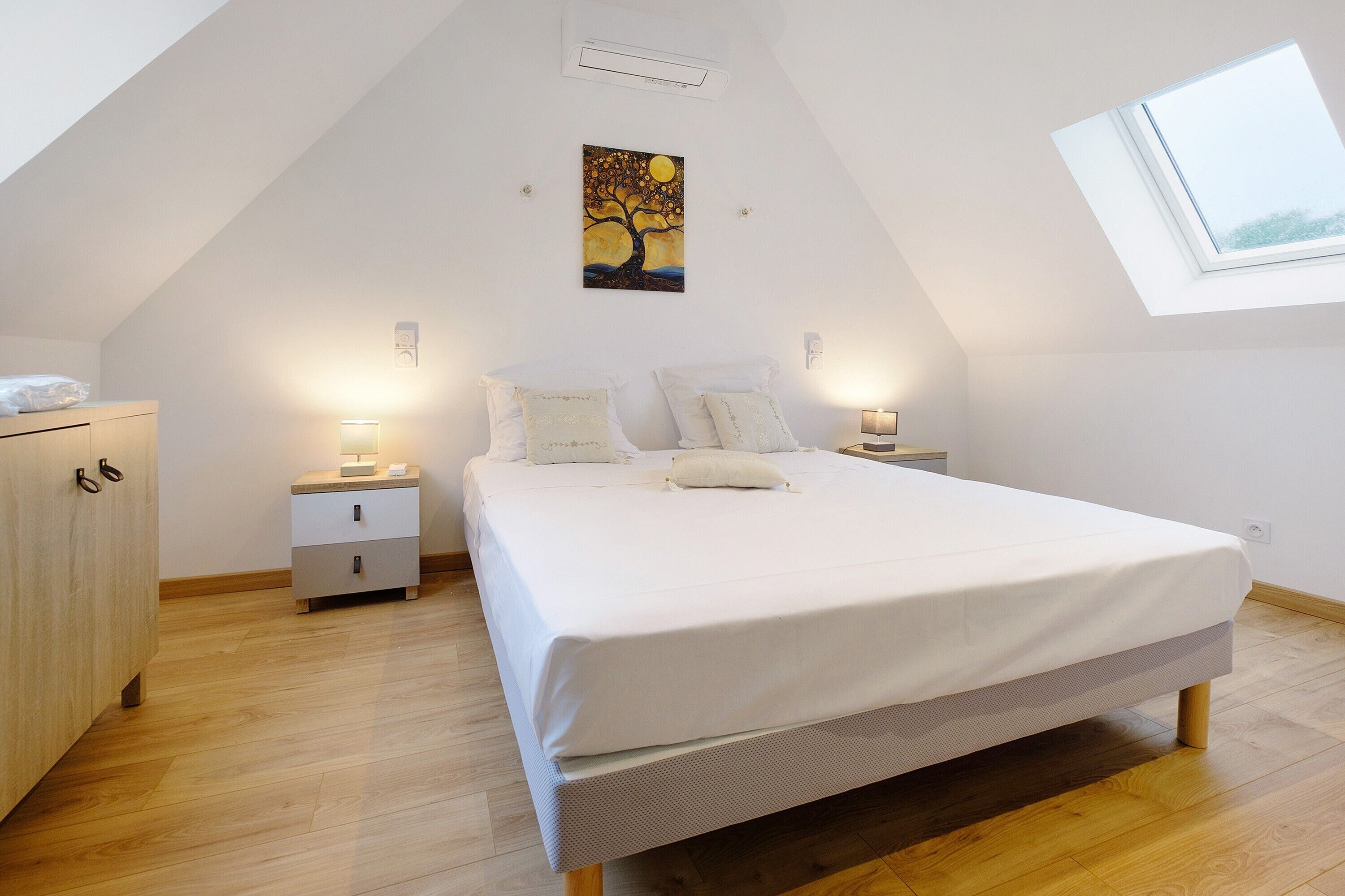 Een slaapkamer met schuine wanden, een bed met wit beddengoed, twee nachtkastjes met lampen, een houten kledingkast en een dakraam.
