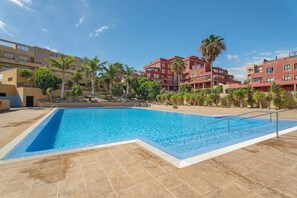 Una piscina con suelo de baldosas, rodeada de edificios de varios pisos con balcones, palmeras y un cielo despejado.
