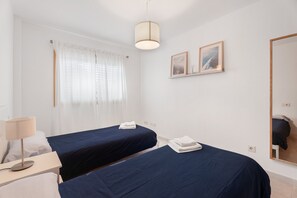 Una habitación de hotel con dos camas, una mesita de noche con lámpara, una ventana con cortinas y un espejo colocado en la pared.