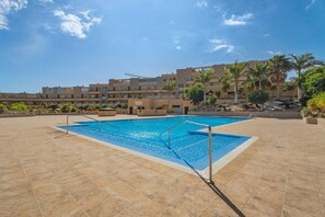 Una piscina con solárium empedrado, rodeada de palmeras y un edificio de varios pisos al fondo.