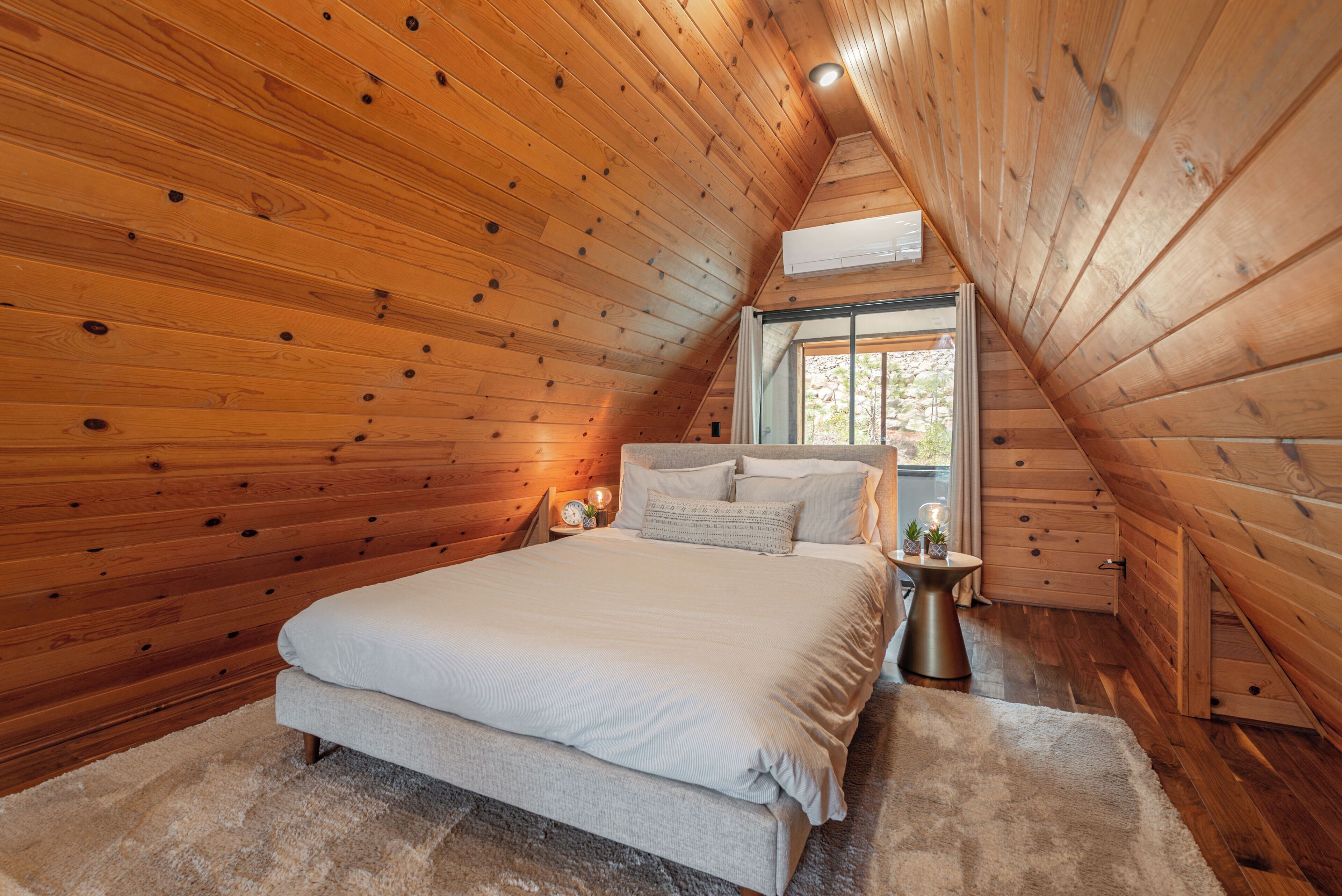 Une chambre avec un plafond en pente en bois, un grand lit, une table de chevet et une fenêtre donnant sur les arbres.