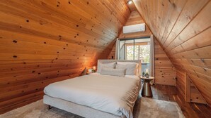 Un dormitorio con techo inclinado de madera, una cama grande, una mesita de noche y un ventanal con vistas a árboles.