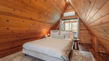 Une chambre avec un plafond en pente en bois, un grand lit, une table de chevet et une fenêtre donnant sur les arbres.