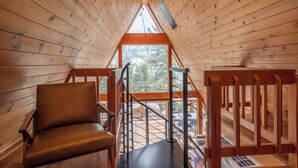 Interior de una cabaña de madera con techo inclinado, una silla de madera y vistas a través de un ventanal grande.