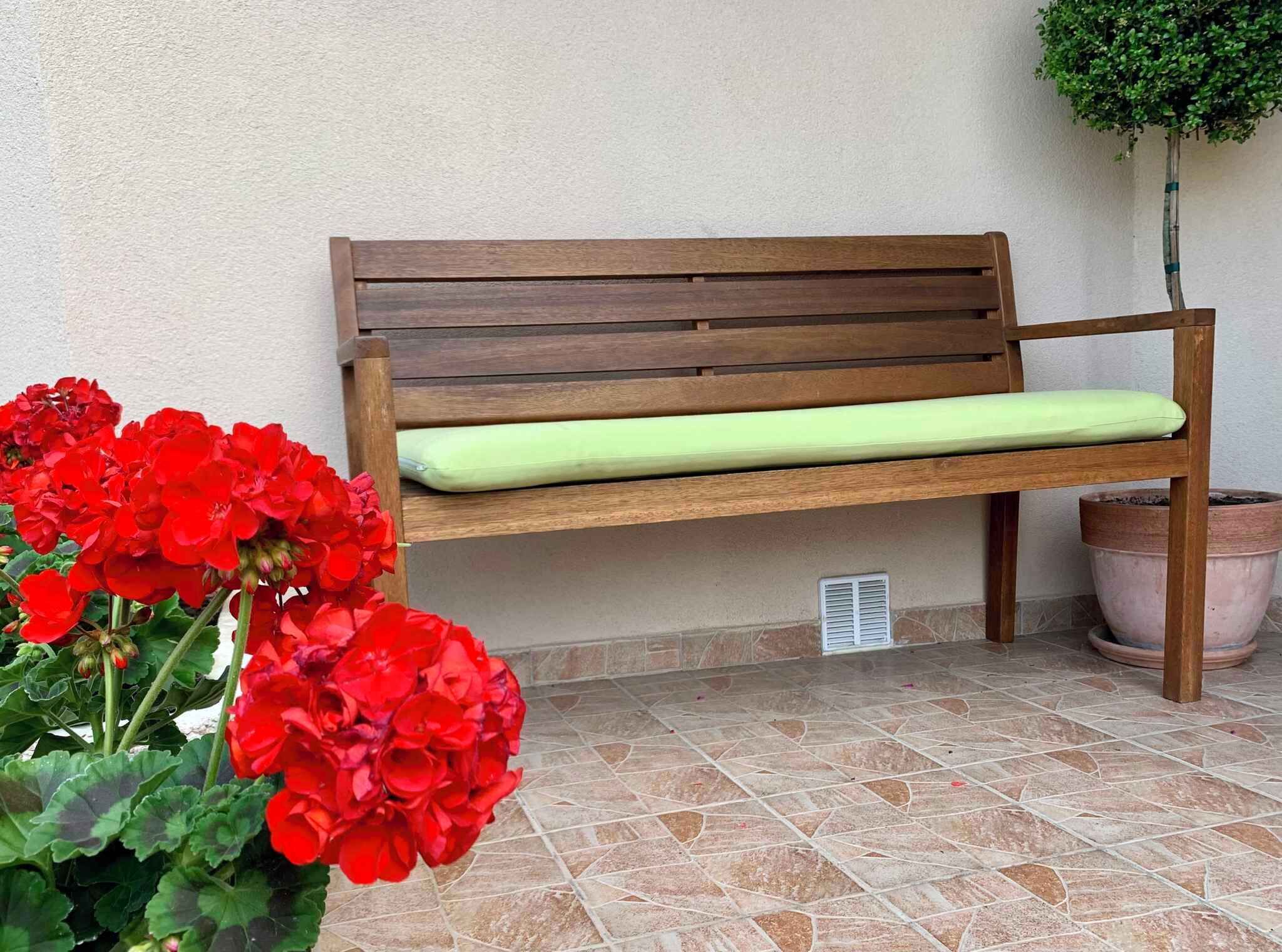 Una panca di legno con un cuscino verde, una pianta in vaso e fiori rossi su un pavimento piastrellato.