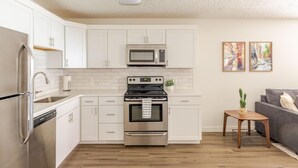 A modern kitchen with white cabinets, stainless steel appliances, and a wooden floor. There is a small side table with a plant and a framed picture on the wall. - Labor Day Getaway | Pool, Spa & Pickleball (St. George)