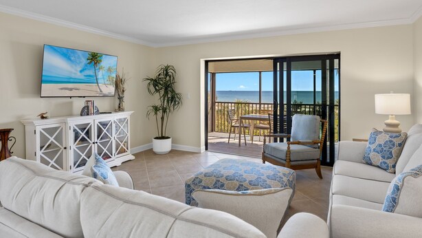 A living room with a large TV, a white cabinet, a potted plant, a balcony with a table and chairs, and a view of the ocean.