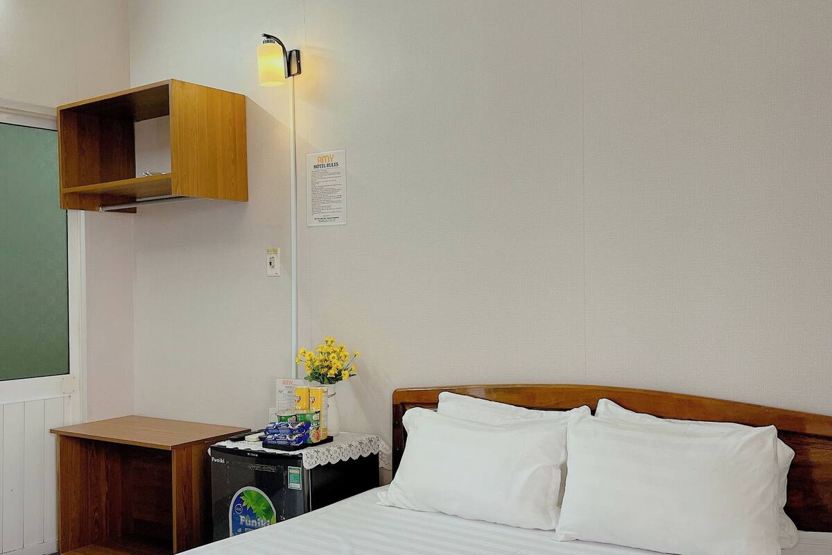 A hotel room with a bed, a nightstand, a wall-mounted air conditioner, and a wooden shelf.