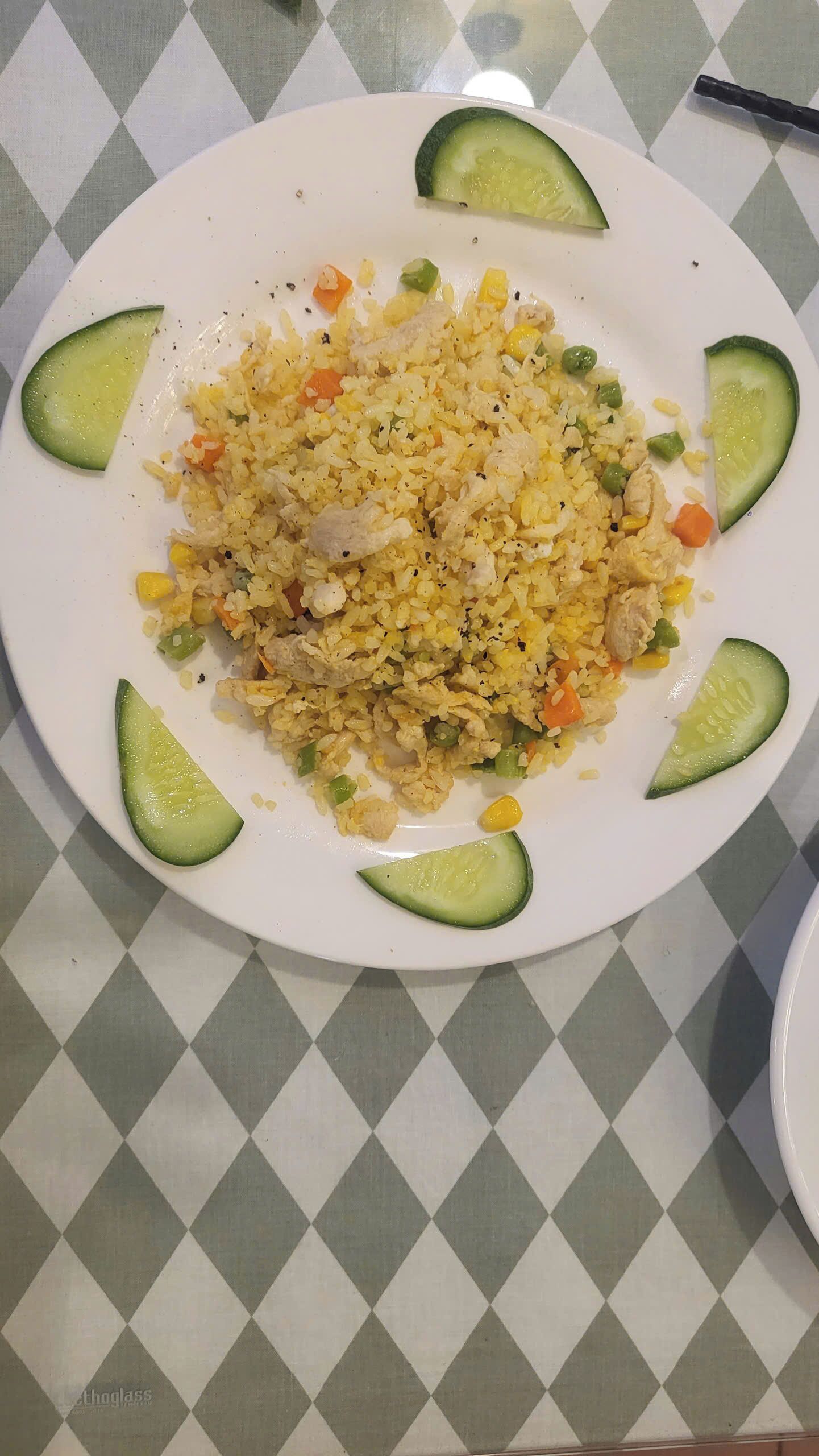 A plate of fried rice with vegetables and chicken, surrounded by cucumber slices on a table with a checkered tablecloth.