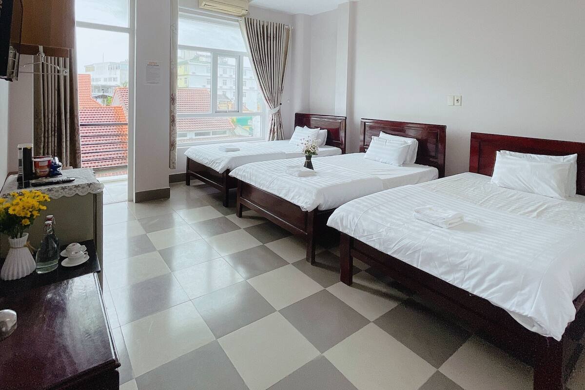 A hotel room with two single beds, a desk with a vase of flowers, a window with curtains, and a view of buildings outside.