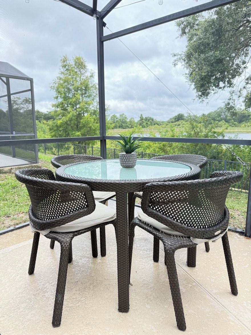 Een ronde glazen tafel met vier rieten stoelen, een potplant en een constructie van metaal.