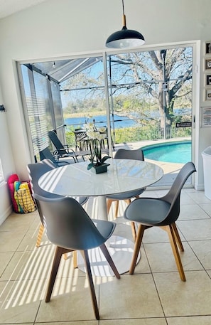 Uma área de refeições com uma mesa redonda e cadeiras, um lustre pendente e vista para uma piscina e árvores através de portas de vidro.