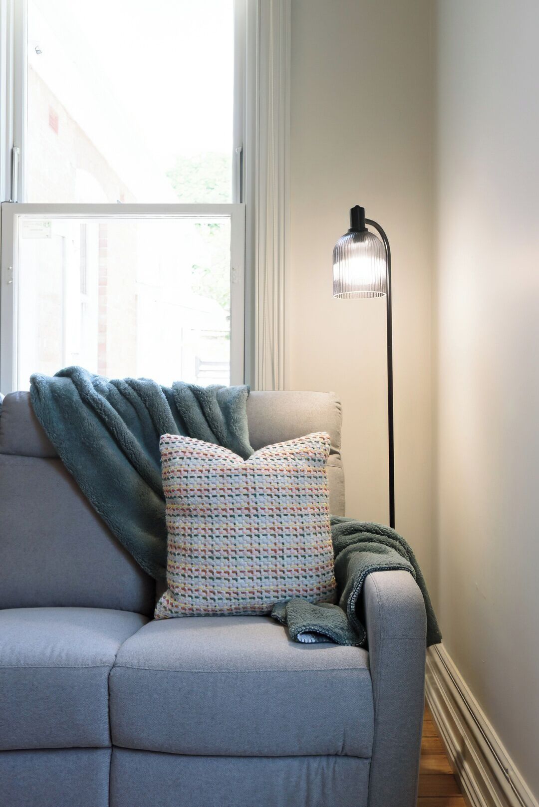 A grey sofa with a patterned pillow and a throw blanket, a floor lamp, and a window with a view of a building outside.