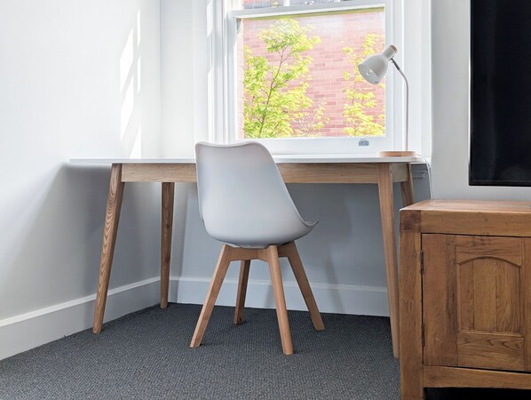 Un espacio de trabajo con escritorio, silla, lámpara y vista a un edificio de ladrillo a través de una ventana.