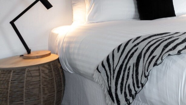 Una cama bien tendida con sábanas blancas y un plaid estampado con diseño de cebra, una lámpara negra sobre una mesita de noche de madera redonda, y un cuadro abstracto minimalista en la pared.