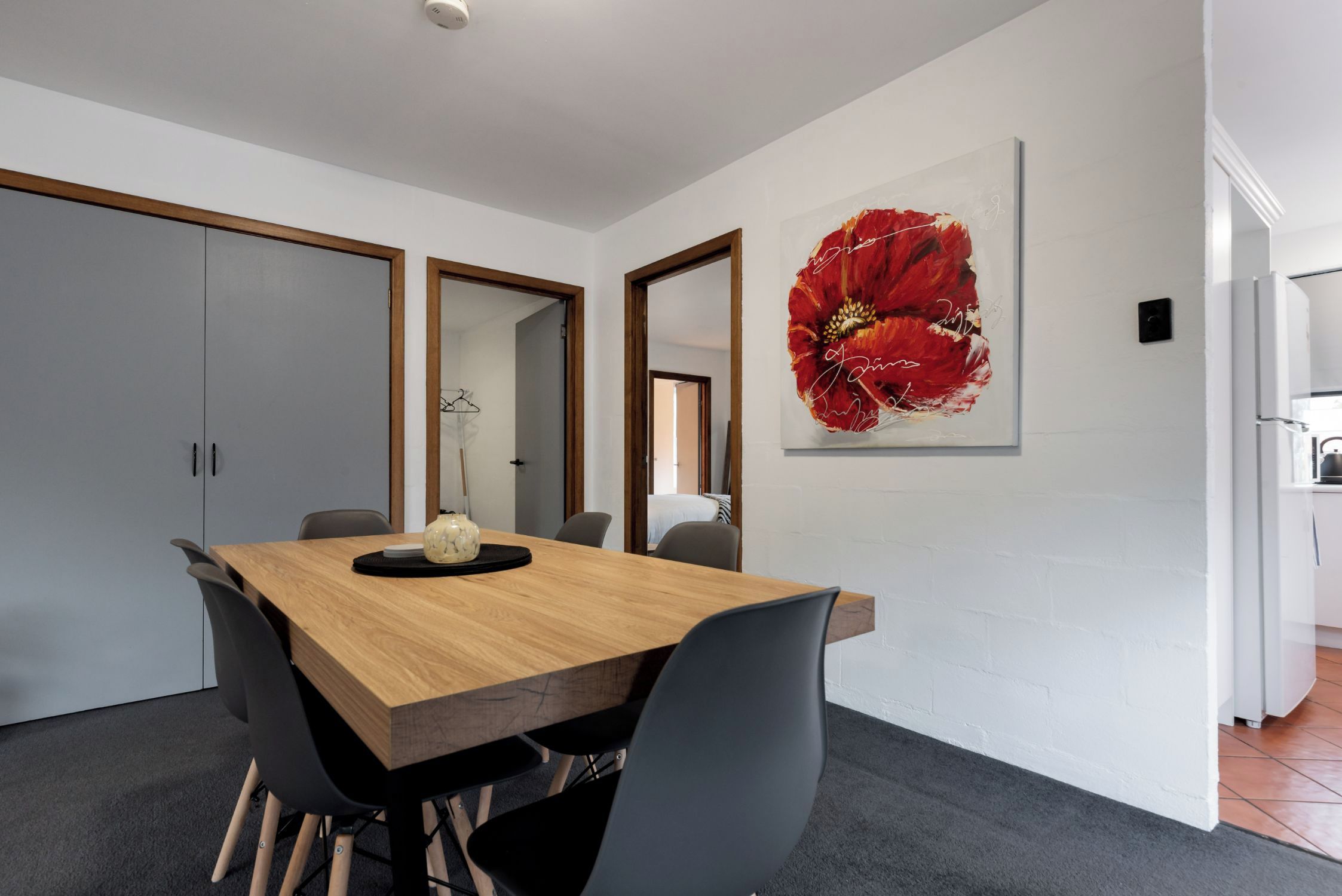 A modern dining area with a wooden table, black chairs, and a large red flower painting on the wall.