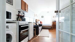 A compact kitchen with a washing machine, oven, and microwave. There is a bed with a headboard and a bedside table with a lamp.