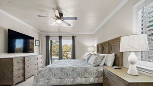 A bedroom with a bed, a ceiling fan, a TV, a window with curtains, and a nightstand with a lamp.