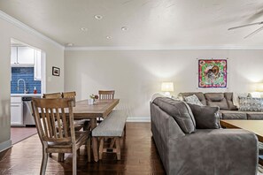 Ein Wohnzimmer mit grauem Sofa, Esstisch aus Holz und Stühlen. Im Hintergrund ist eine Küche mit blauen Fliesen zu sehen.
