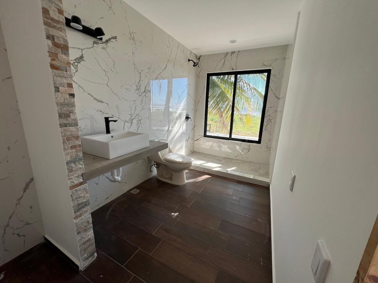 Une salle de bain avec un mur en marbre, un lavabo blanc, des toilettes et une grande fenêtre donnant sur un paysage verdoyant.