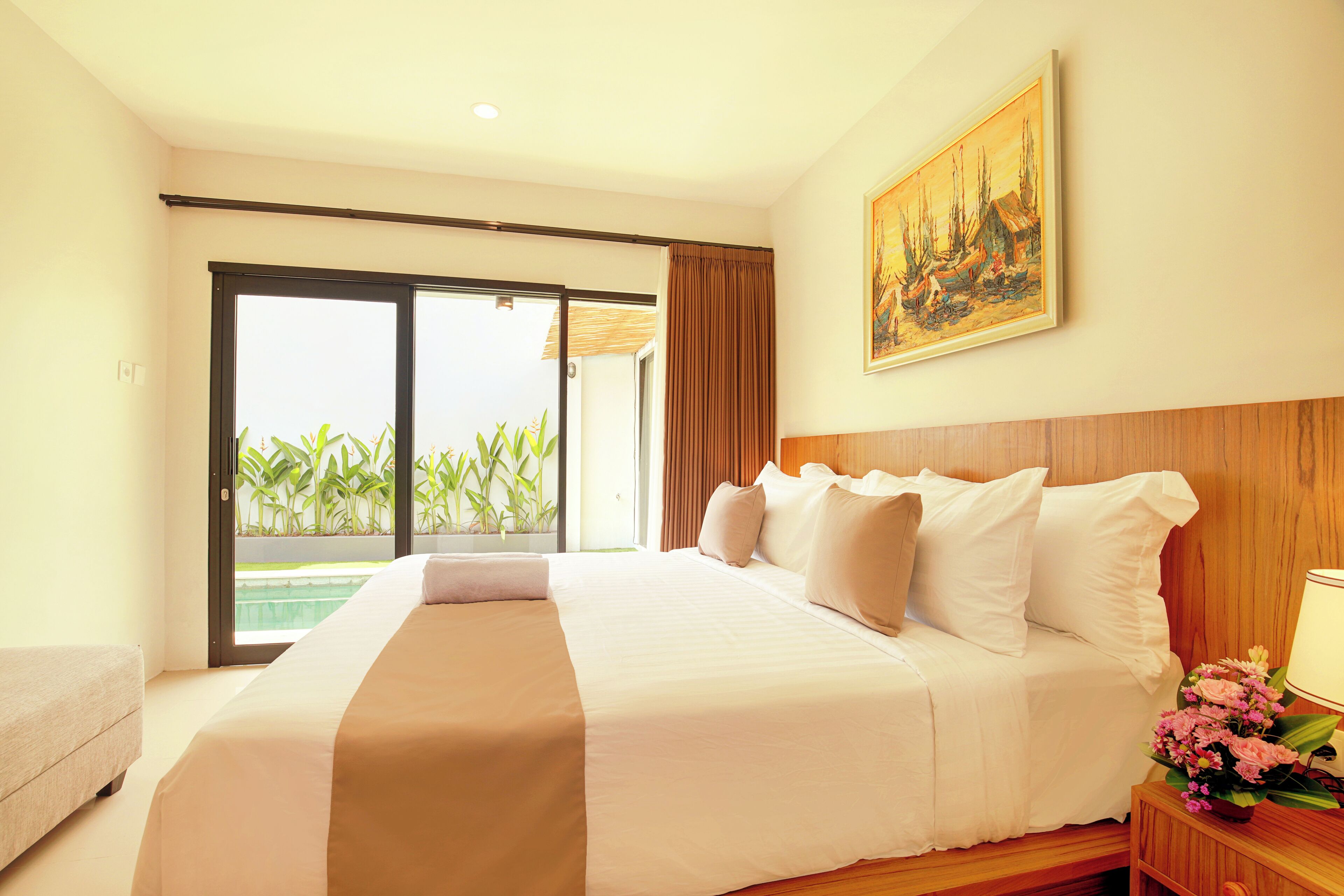 Une chambre d’hôtel avec un grand lit, une tête de lit en bois, un tableau au mur et une vue sur la piscine, visible à travers la porte-fenêtre coulissante.