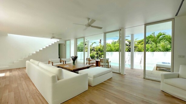 A modern living room with white sofas, a wooden coffee table, and a dining area with chairs and a table. There is a staircase leading to an upper level, and large sliding glass doors open to a balcony with a view of greenery and a pool. - Sunset Bungalow (Canggu)