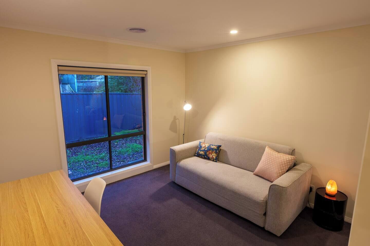 A room with a beige sofa, a wooden table, and a large window with a view of a backyard.
